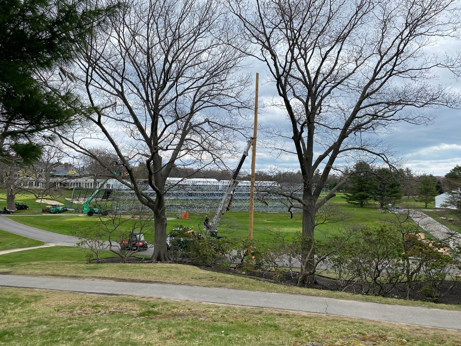 During: Digger derrick raises a wood pole while grandstands go up across the course. Golf Course Netting at The Country Club, Brookline
